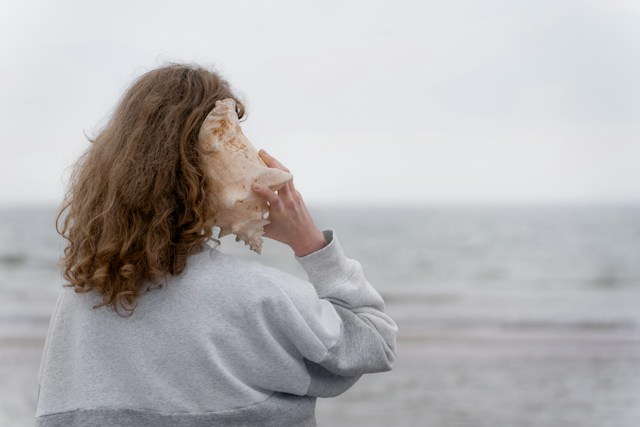 Personne tenant un coquillage contre son oreille, face à la mer